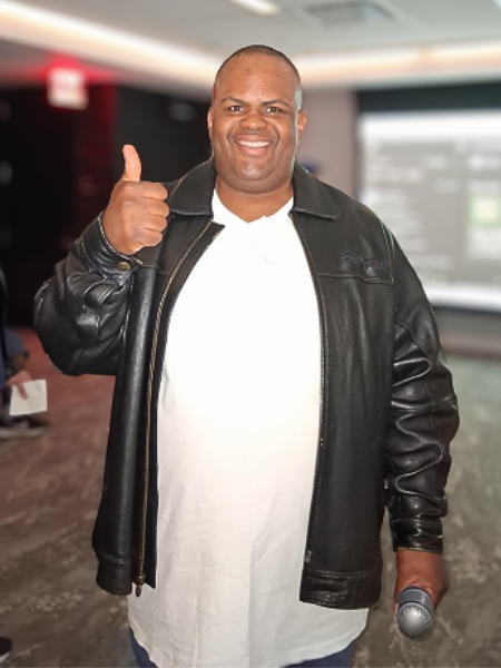 Kenneth with his thumb up and holding a mic. wearing a white t-shirt and black leather jacket