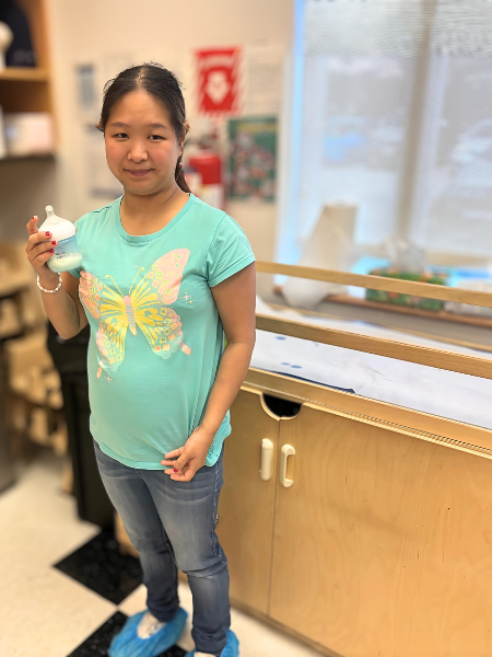 Mei-Feng stands with a bottle of infant formula in her hand, you can see a changing table in the background