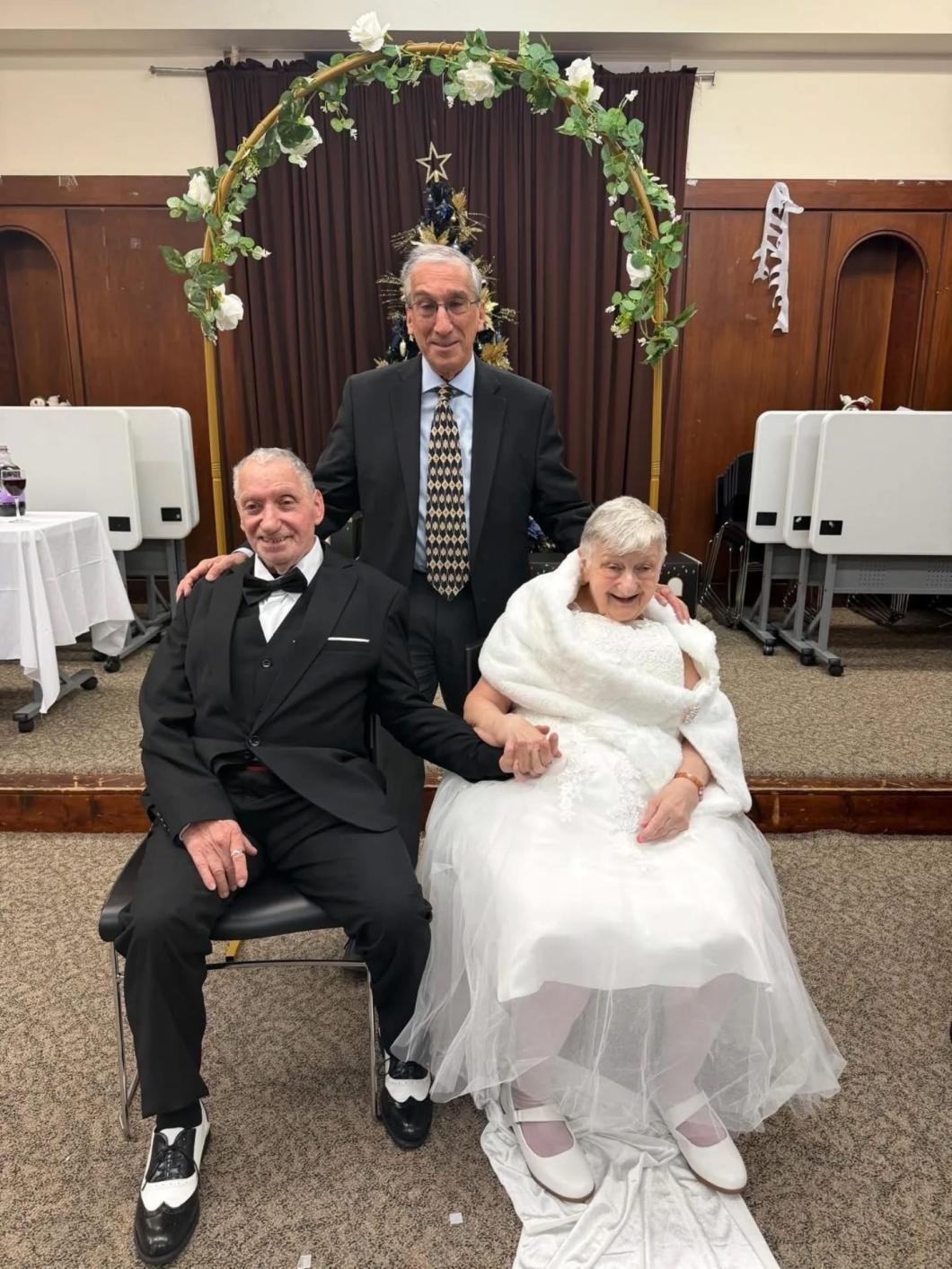 Melvin and Joan in formal attire seated and holding hands, with Rabbi Jonathan Pearl standing behind them under a decorated arch