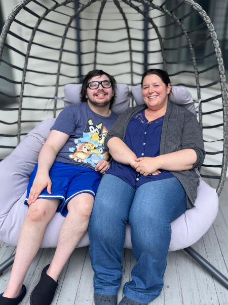 Tyler sits in a hanging chair with a staff member, they smile for the photo