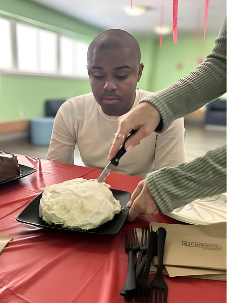 Justin sits at a table at the Kingston center, someone is cutting a cake in front of him as he watches on.