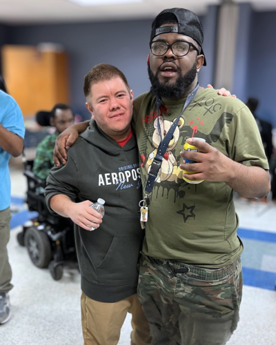 two men stand together. one has his arm over the shoulders of the other