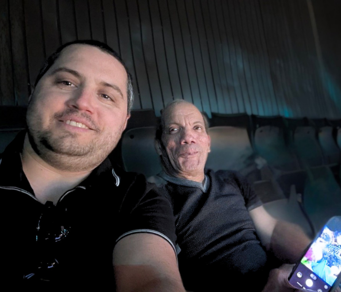 Two men sit at what may be a game, howard is holding his phone so you can see the screen