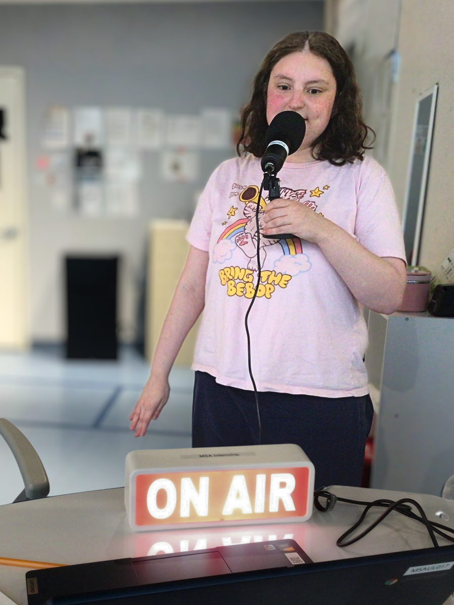 Kali stands holding a mic, in front of her is a laptop and a lit "ON AIR" sign