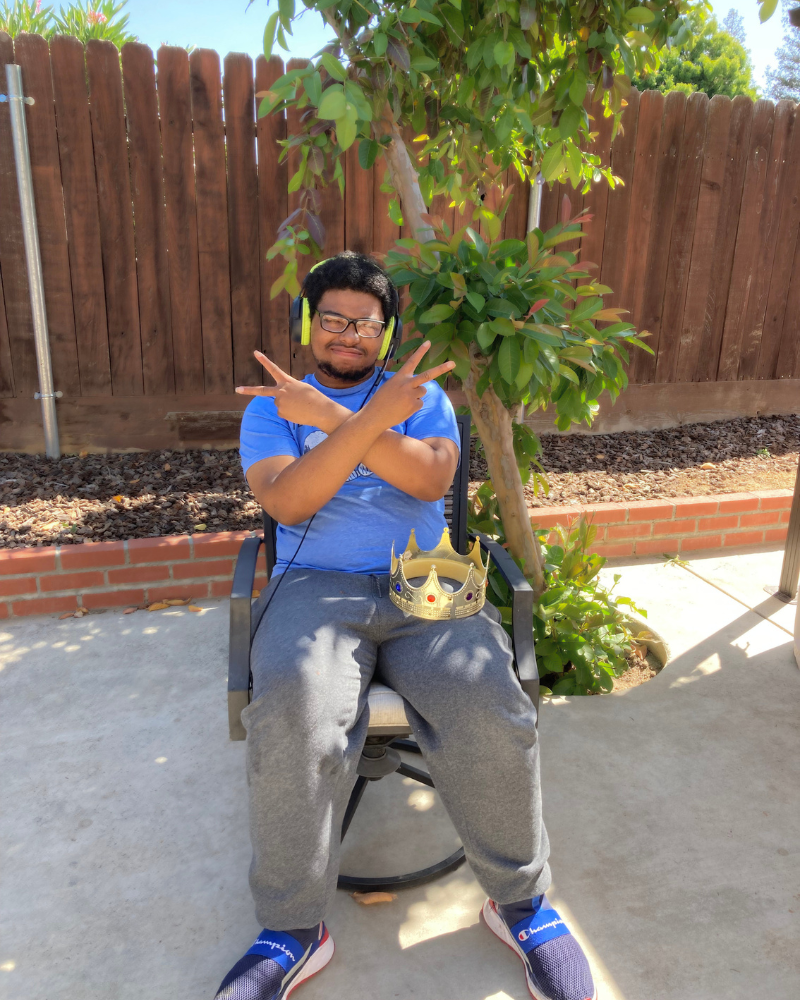 Jabrell sitting on a chair outdoors, with a crown on their lap, making a crossed arm gesture.