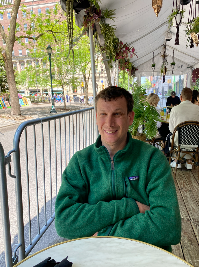 Person in green jacket sitting at outdoor café table with greenery and city street in background