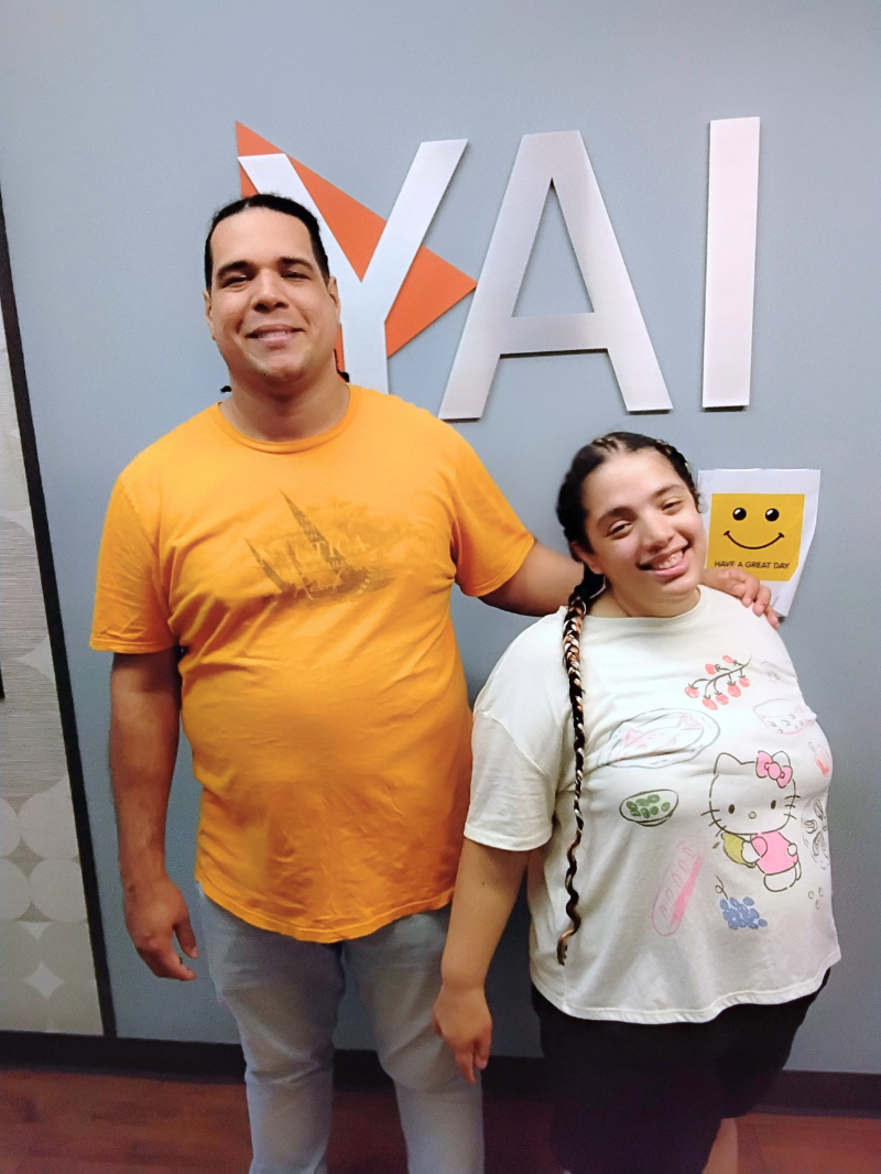 Two people posing in front of a blue wall with large “YAI” sign; one wears an orange shirt, the other wears a white patterned top and smiles while standing close together.