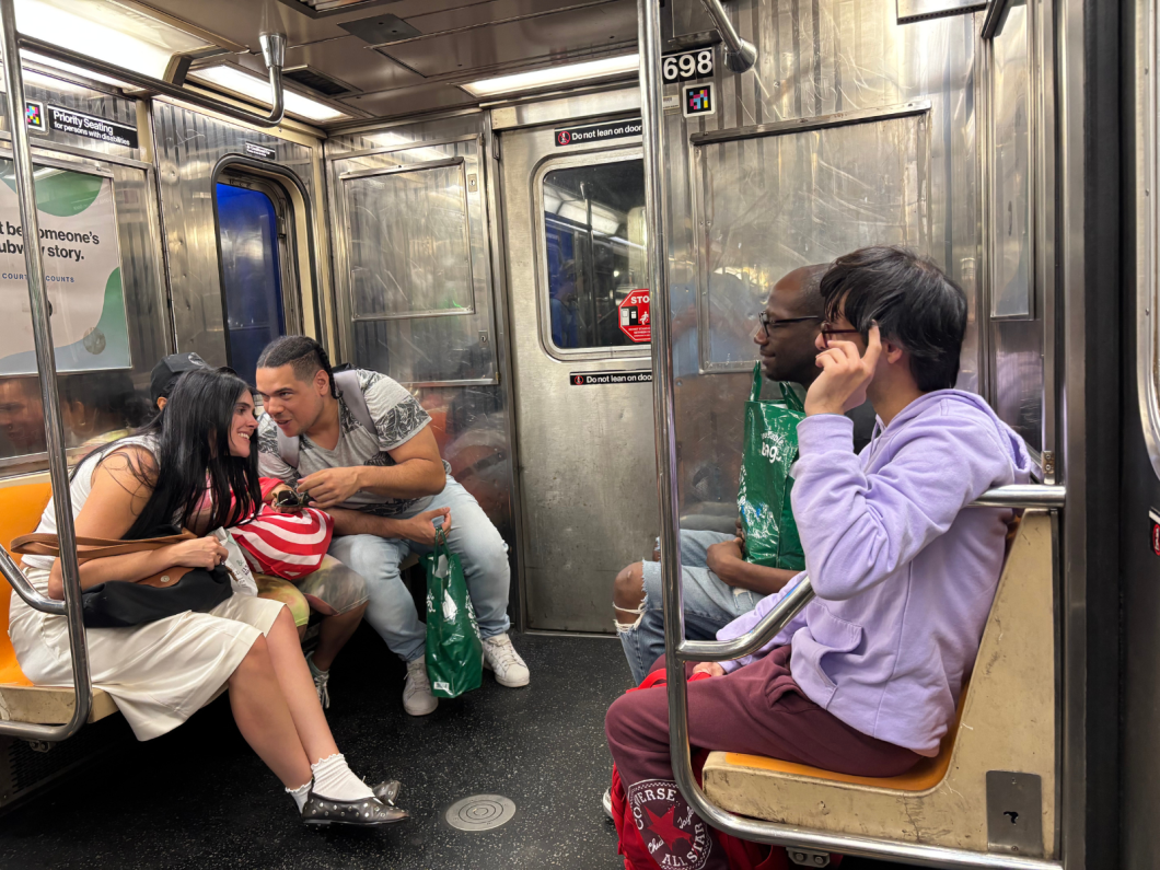 Inside a subway car, several passengers sit; one person in a purple hoodie rests a hand near the face while others chat and look toward the windows.