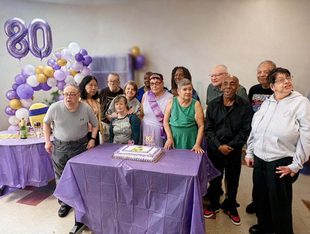 Group of people from the day hab gathered at Veronica's birthday celebration around a purple table with a decorated cake, standing in a room decorated with purple and gold balloons and a large balloon sculpture behind them.