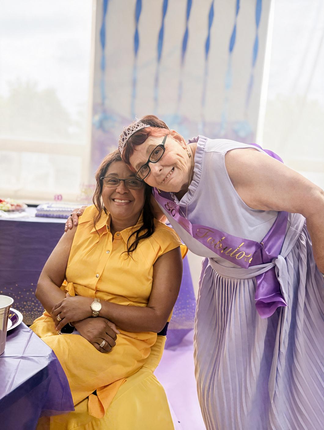 Two people smiling at Veronica's birthday celebration, one in a yellow dress and glasses seated at a table, the other in a light purple dress leaning in with an arm around them.