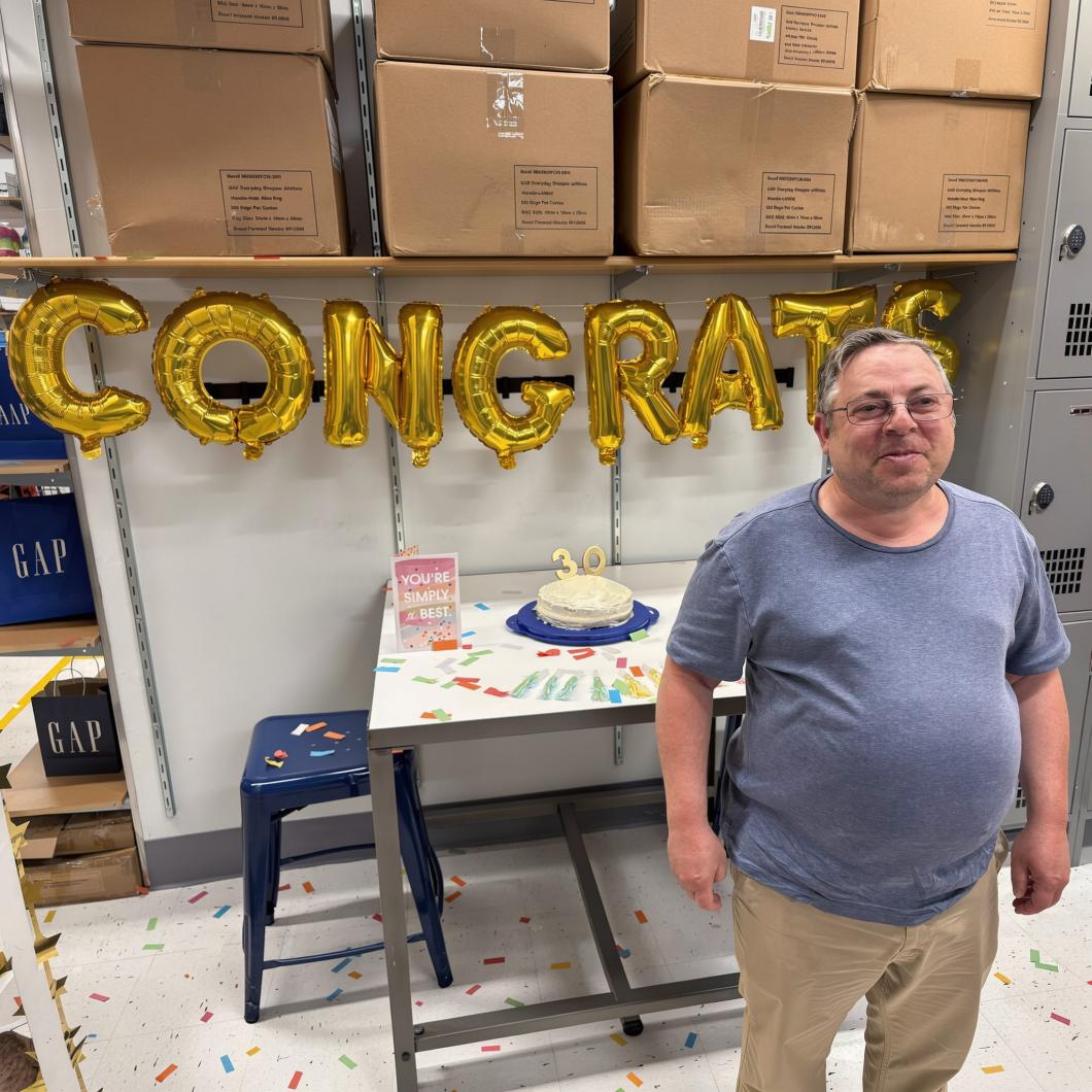 Man stands in staff office at GAP, a cake and gold "CONGRATS" balloons are behind him. 