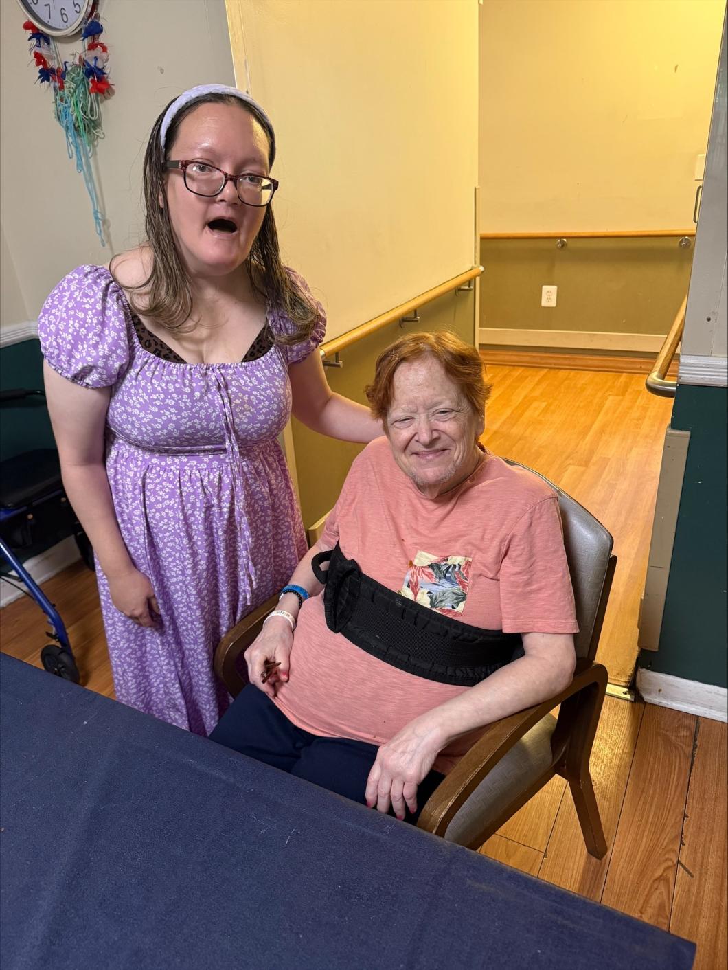 Young woman in purple dress stands beside seated older woman in chair at the Forest Hills residence